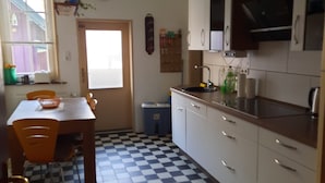 Een keuken met een zwart-witte betegelde vloer, een eettafel met stoelen, kasten en een raam met jaloezieën.