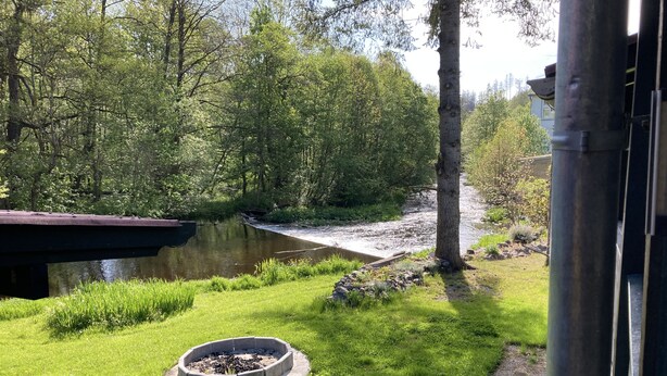 Een tuin aan de rivier met een ronde vuurkuil, een bank en uitzicht op een rivier en bomen.