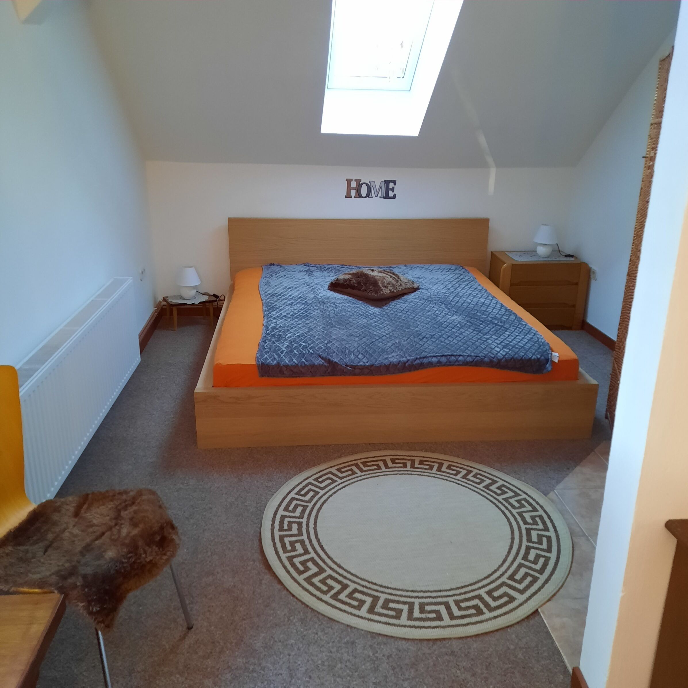 Une chambre avec une lucarne, un lit doté d’un matelas orange, une tête de lit, une table de chevet avec une lampe, une petite chaise et un tapis décoratif.