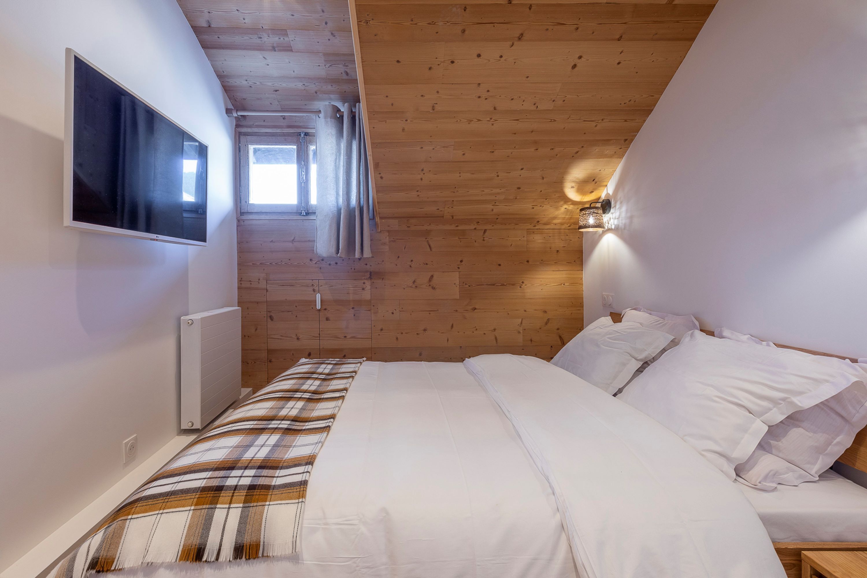 Un dormitorio con cama, ropa de cama blanca, un televisor montado en la pared y techo de madera.