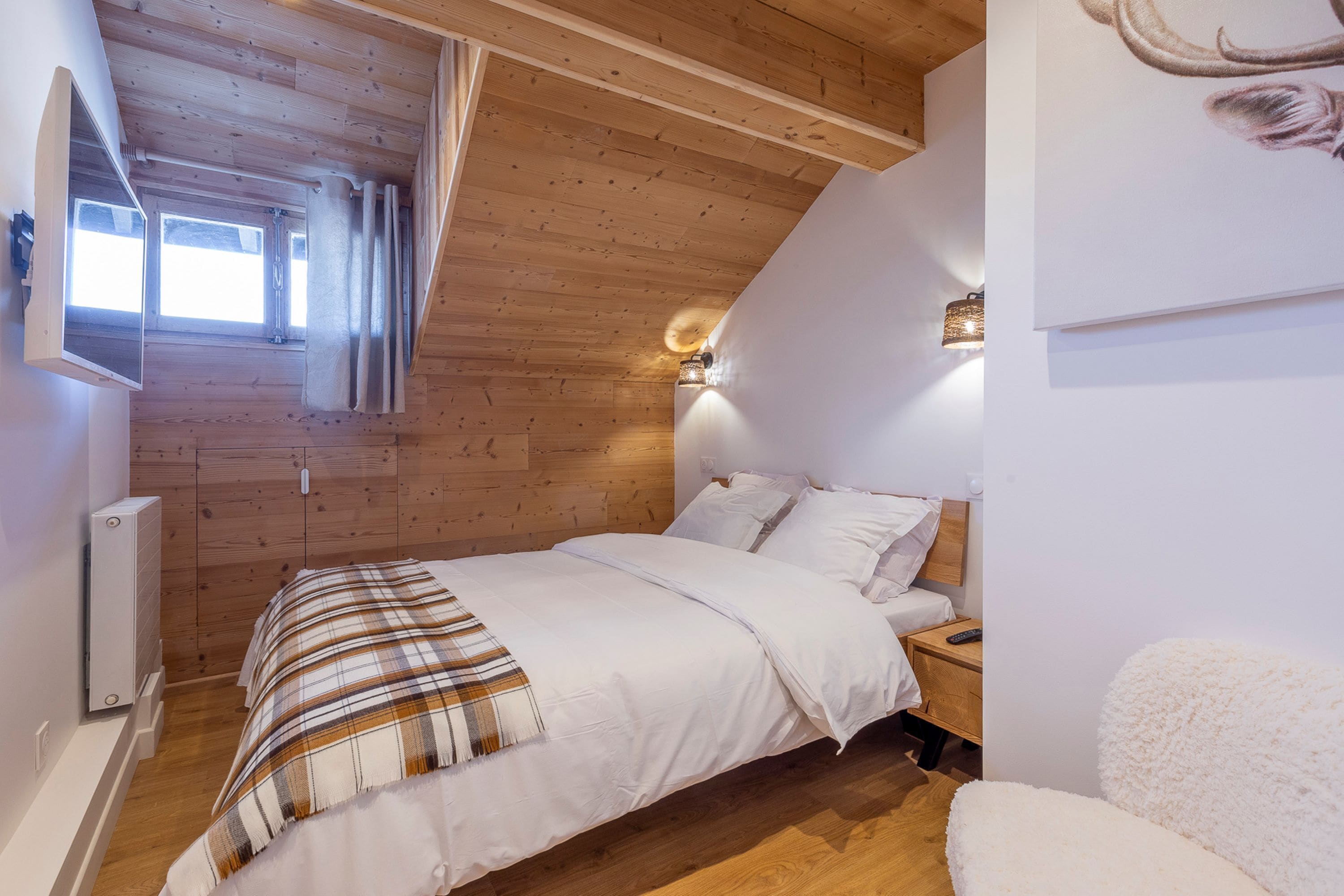 A bedroom with a wooden ceiling, a bed with white bedding, a nightstand, a wall-mounted light, and a window with curtains.