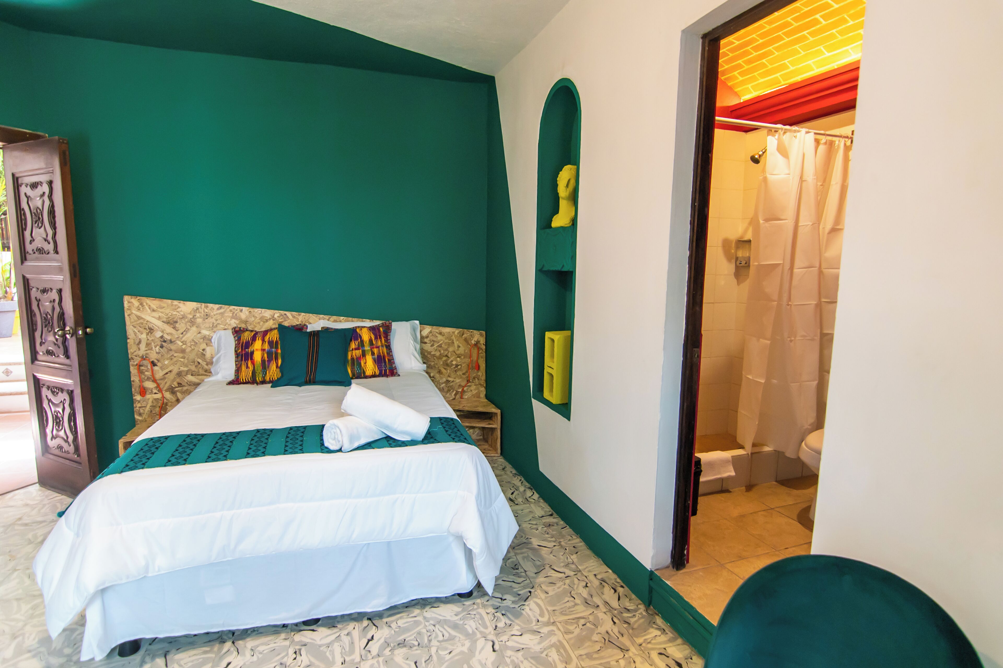 A bedroom with a bed, a wooden headboard, a green wall, and a bathroom visible through an open door.