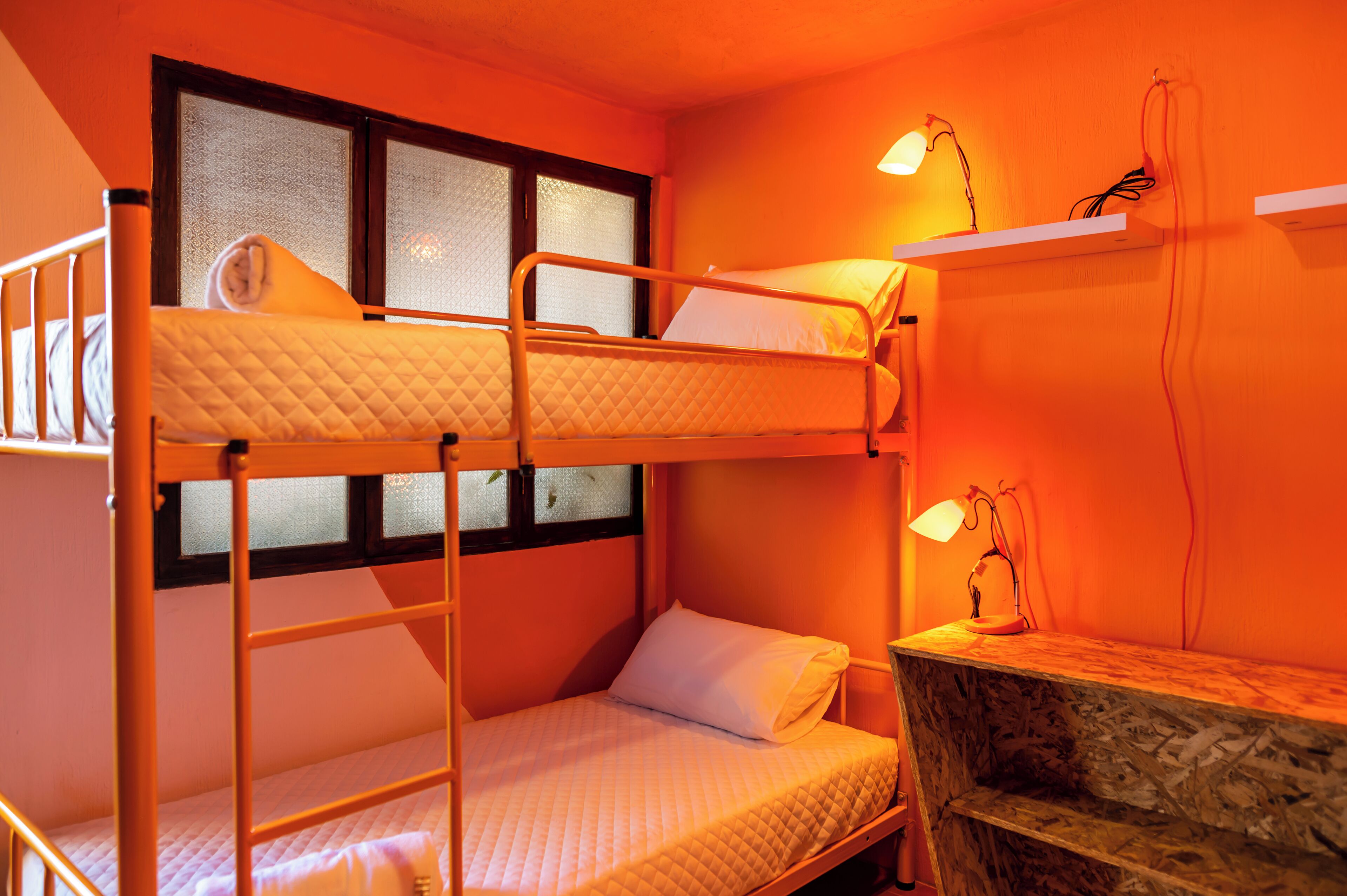 A bunk bed room with a window, a desk with a lamp, and a shelf with electrical cords.