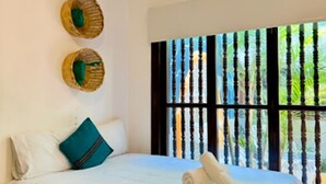 Una cama bien hecha con sábanas blancas y un cojín azul verdoso, una ventana con persianas verticales y un adorno de pared tejido.