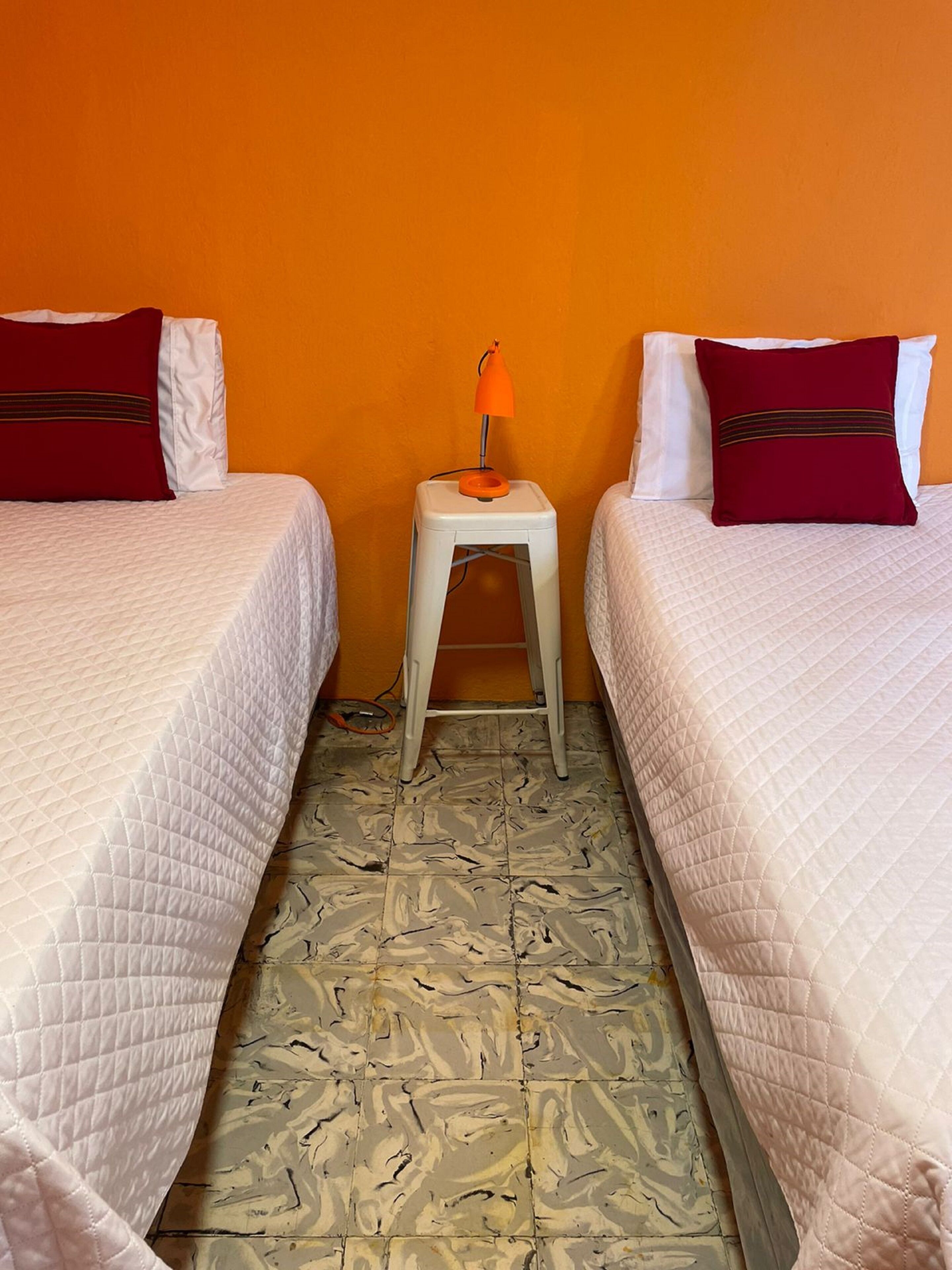 Quarto de hotel com duas camas, cada uma com colcha branca acolchoada e travesseiro vermelho. Entre as camas, há uma mesinha de cabeceira branca com uma luminária laranja. O chão possui um design de piso em mosaico.