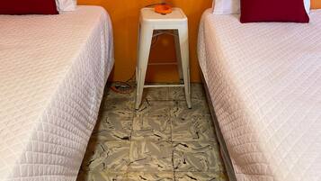 Quarto de hotel com duas camas, cada uma com colcha branca acolchoada e travesseiro vermelho. Entre as camas, há uma mesinha de cabeceira branca com uma luminária laranja. O chão possui um design de piso em mosaico.