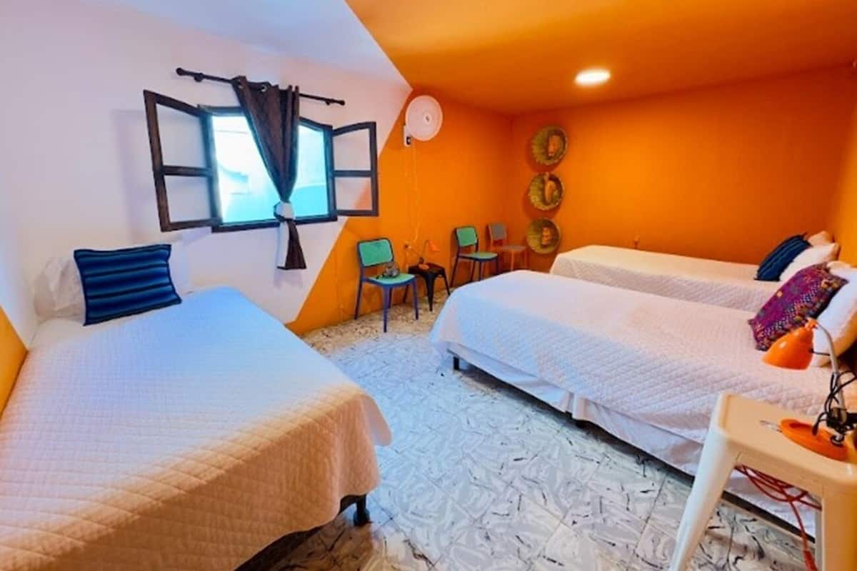 A room with two beds, a window with curtains, a fan, and a small table with a lamp.