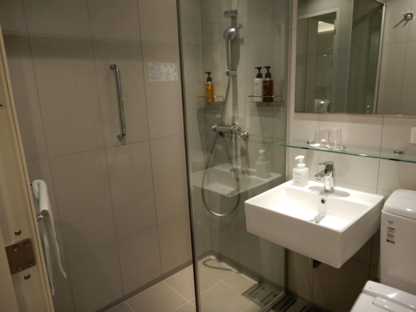 A modern bathroom with a glass-enclosed shower, a white sink with a mirror above it, and a glass shelf with toiletries.