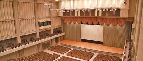 A large auditorium with tiered seating, a stage with curtains, and a ceiling with recessed lighting.