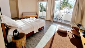 A hotel room with a bed, a wooden side table, a small round ottoman, a balcony with a view of palm trees and the sea, and a chair. - Lea Spa Hotel (Maafushi)