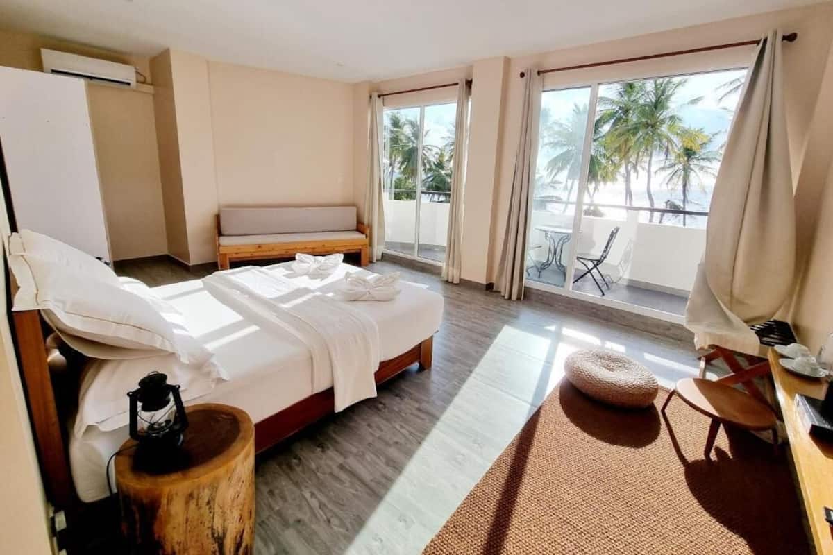 A hotel room with a bed, a wooden side table, a small round ottoman, a balcony with a view of palm trees and the sea, and a chair.