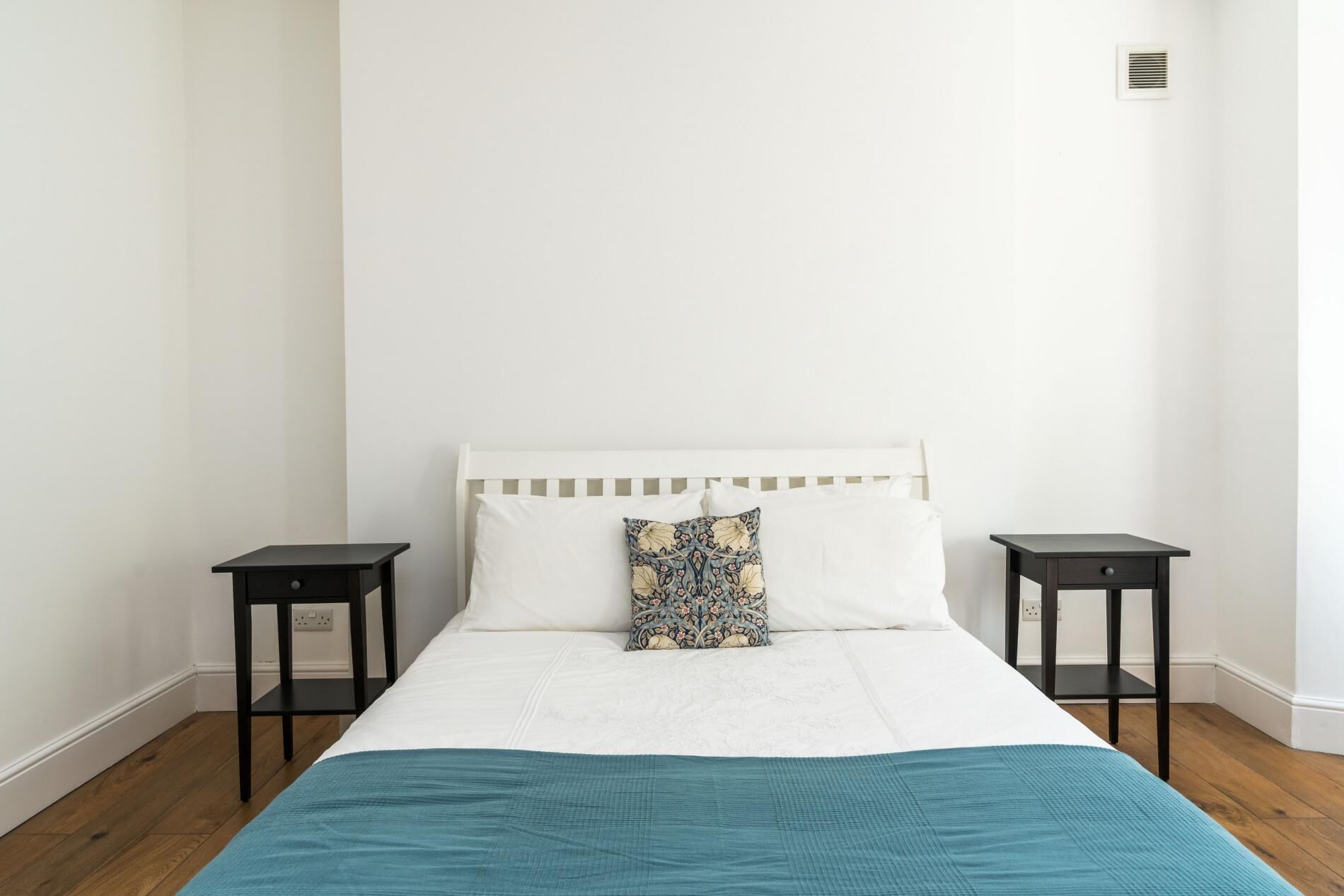 A simple, clean bedroom with a white bed, a teal blanket, two black nightstands, and a patterned pillow.
