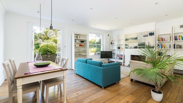 Een ruime woonkamer met houten vloer, een blauwe bank, een eettafel met stoelen en een boekenkast vol boeken.