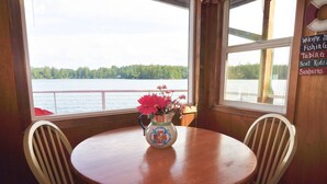 A round wooden table with two white chairs, a vase with pink flowers, and a view of a lake through large windows. - New Listing! Adorable lakefront cabin, private dock, hot tub, gas grill, amazing views (Florence)