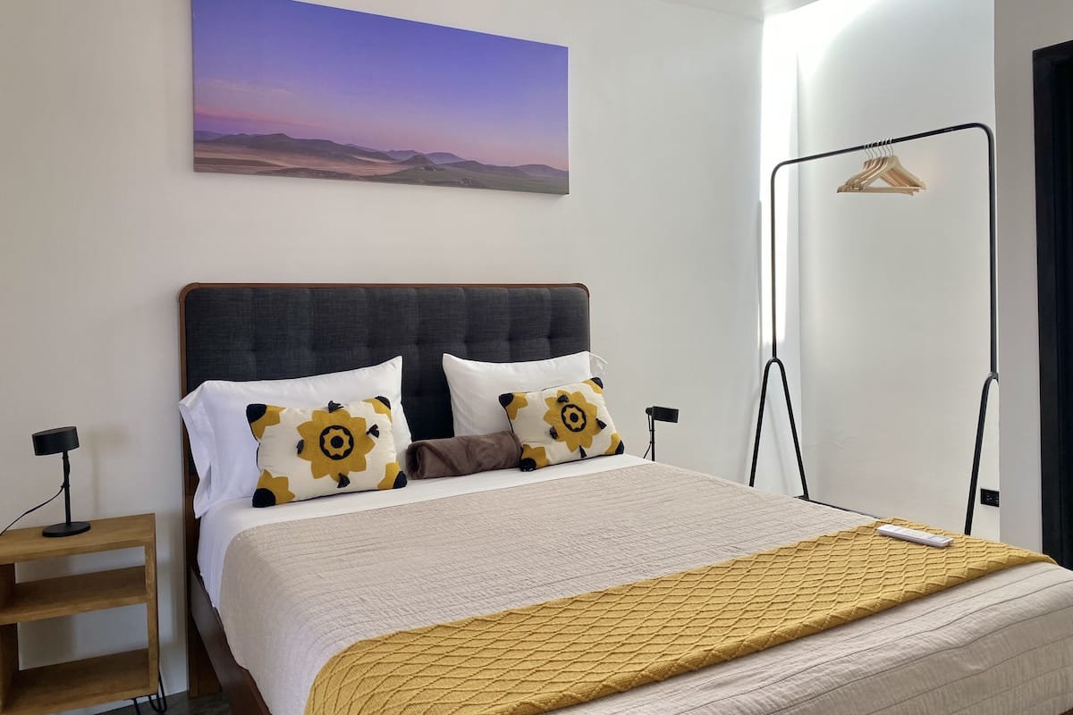 A neatly made bed with a headboard, two pillows with a yellow and black pattern, a bedside table, and a wall-mounted picture of a landscape.