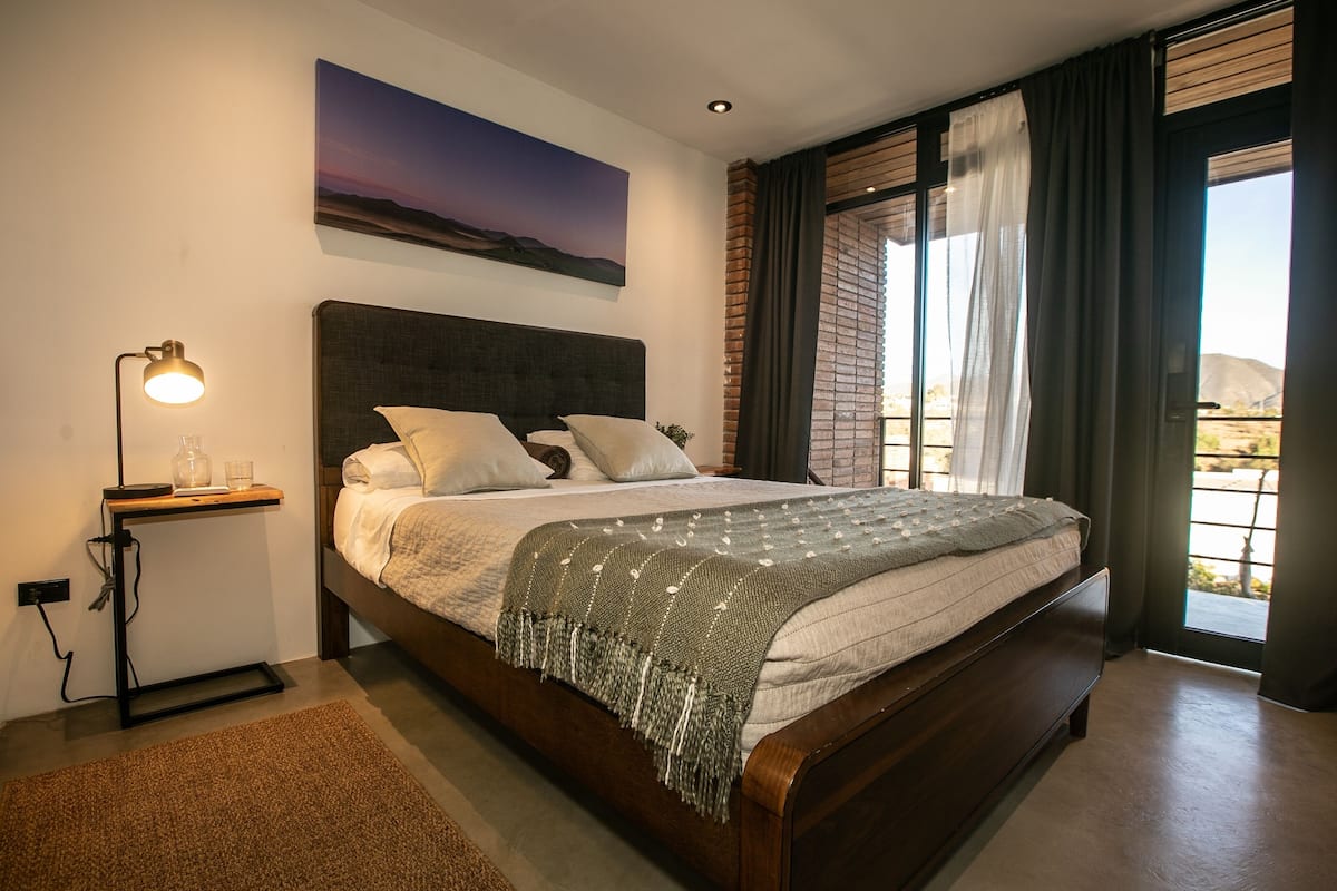 A modern bedroom with a large bed, a bedside table with a lamp, and a view of the outdoors through a window with curtains.