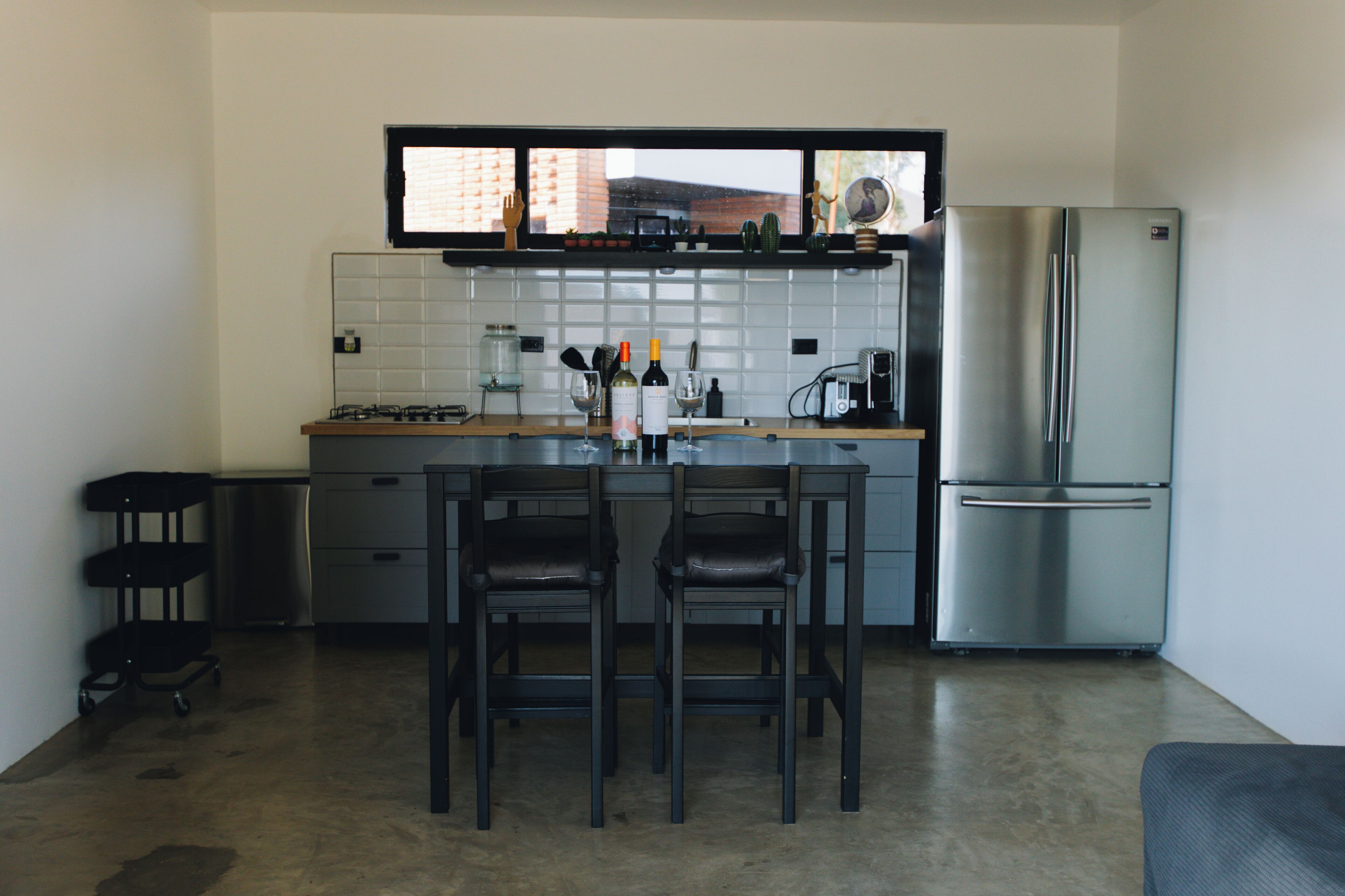 A modern kitchen with a central island, two bar stools, a stainless steel refrigerator, and a window above the counter.