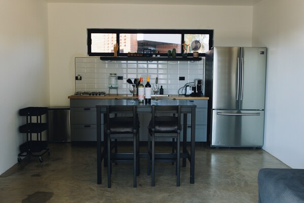 Una cocina moderna con una isla central, dos taburetes de bar, un refrigerador de acero inoxidable y una ventana sobre la encimera.