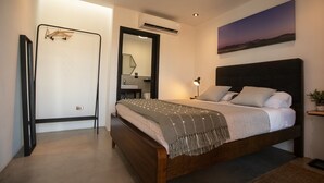 A bedroom with a wooden bed, a nightstand, a wall-mounted air conditioner, a mirror, and a painting of a sunset.