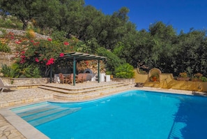 Une piscine agrémentée d’une pergola et d’un espace salon extérieur, le tout entouré d’une végétation luxuriante et de plantes en fleurs.