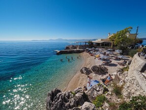 A coastal beach with clear waters, a rocky shoreline, and a building with outdoor seating.