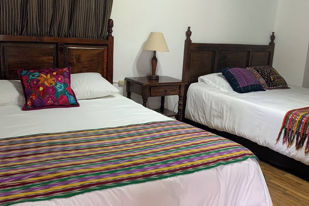 Habitación de hotel con dos camas, cabeceros de madera, una mesita de noche con lámpara y una manta a rayas coloridas.