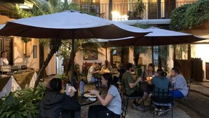 Un espace de restauration en plein air, avec des tables et des chaises placées sous des parasols, entouré de verdure et bordé par un bâtiment doté d’un balcon.