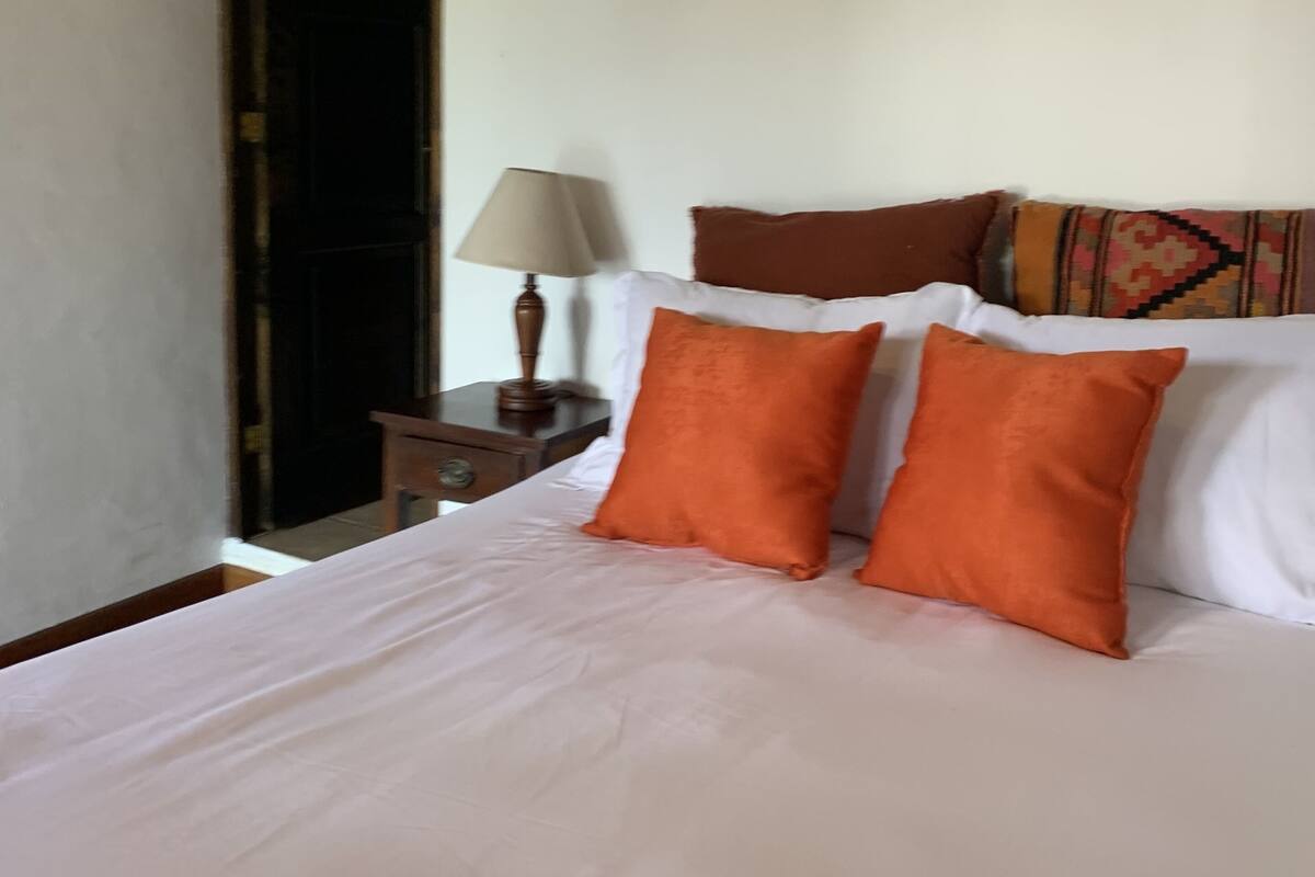 Una cama con almohadas blancas y anaranjadas, una mesita de noche con una lámpara y un plaid estampado.