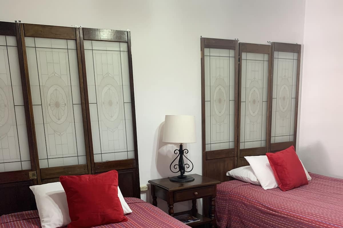 Habitación de hotel con dos camas, cada una con colcha roja y almohadas blancas, una mesita de noche con lámpara y cabecera de madera.