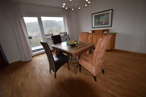 Uma área de refeições com mesa de madeira, duas cadeiras, uma tigela de frutas e vista para o exterior através de uma janela grande.