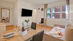 En moderne stue med et fladskærms-tv, et spiseområde med et bord dækket til to personer, og en sofa.