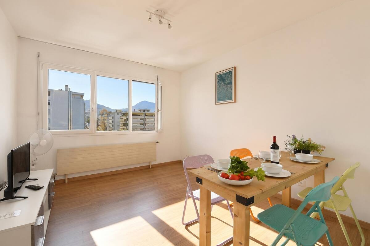 Une pièce moderne et bien éclairée, avec une table à manger dressée pour deux personnes, un bureau et une grande fenêtre donnant sur la ville.