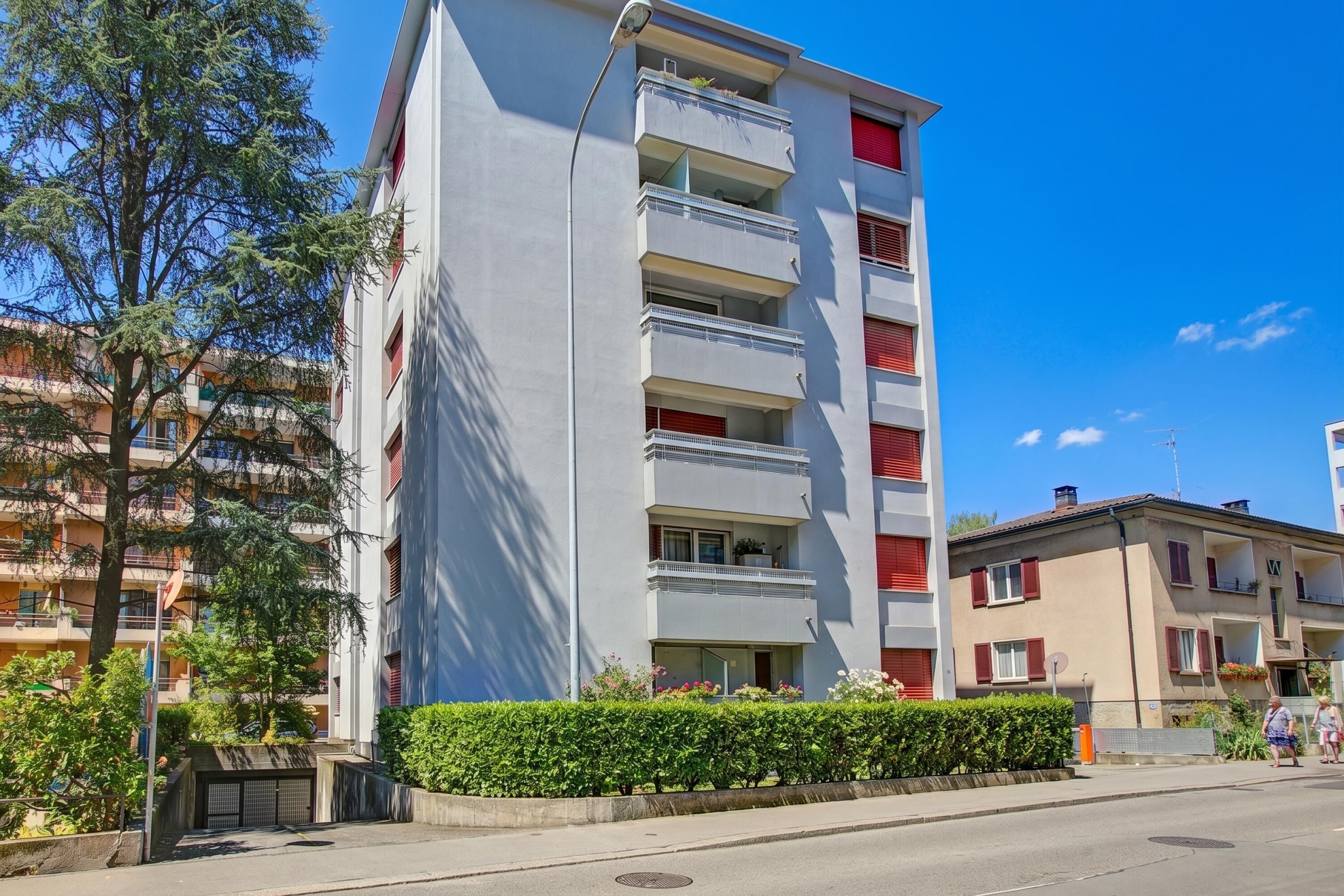 A multi-story residential building with balconies, a clear sky, and a hedge in the foreground.