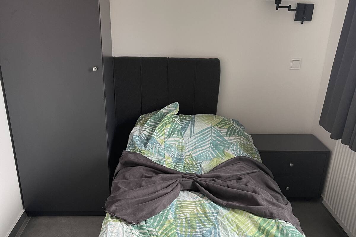 A bed with a leaf-patterned bedspread, a dark headboard, a bedside table, and a wardrobe.