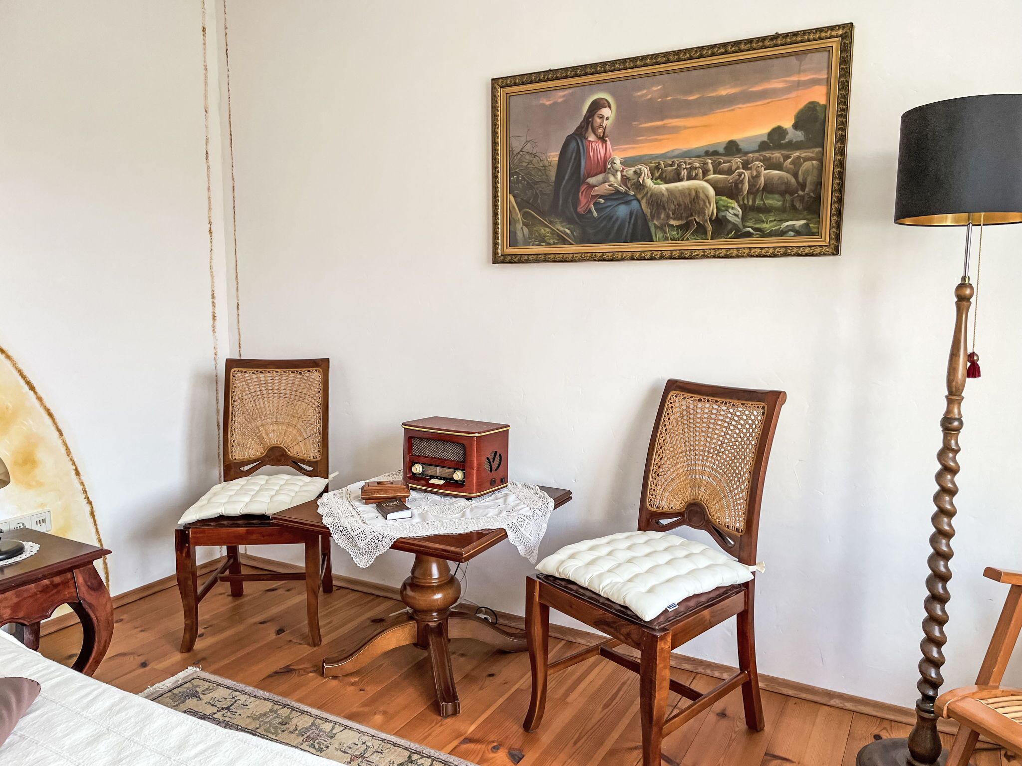 Quarto com duas cadeiras de madeira, uma mesa pequena com toalha de renda, um rádio antigo e um quadro na parede.