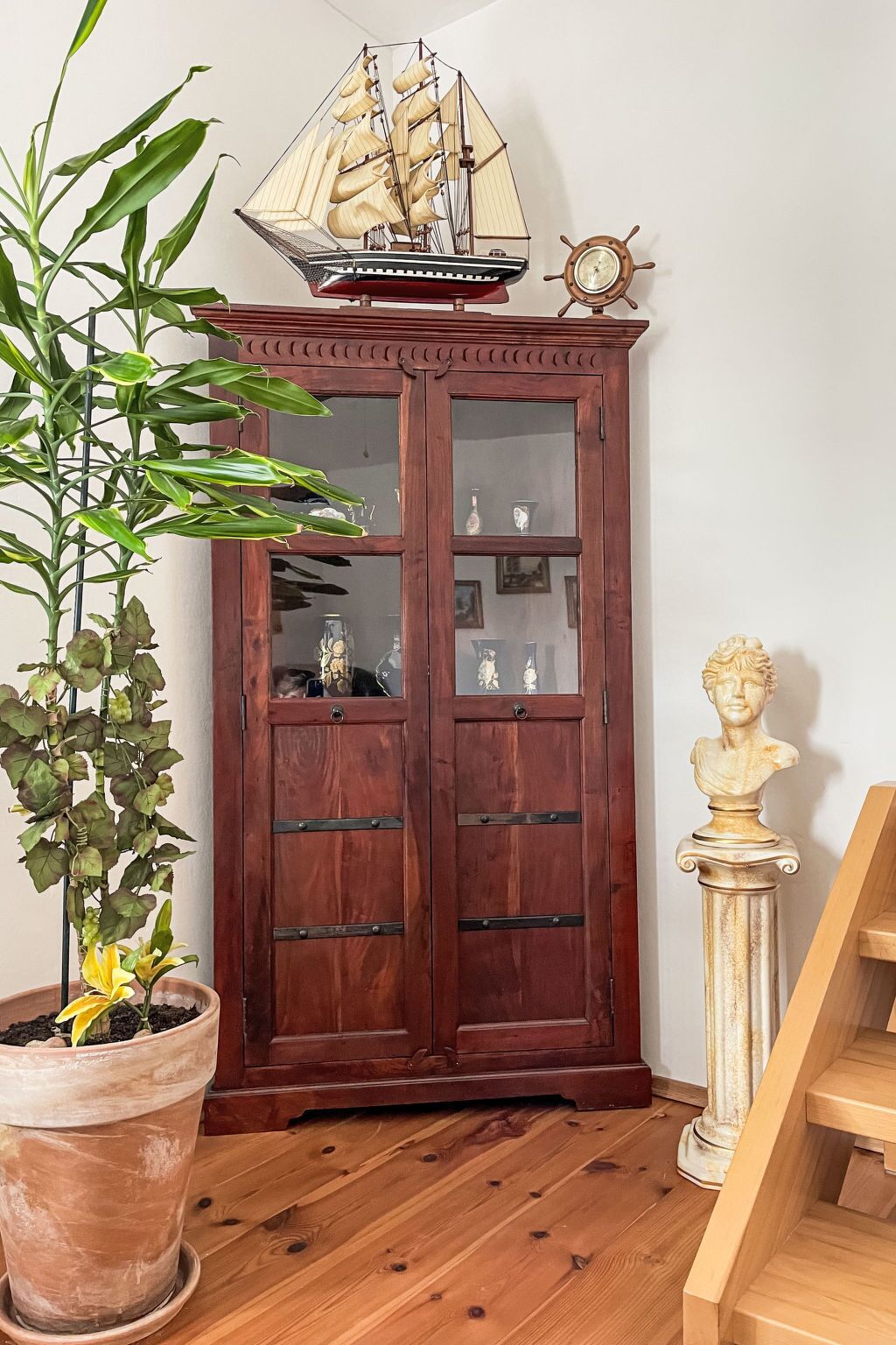 Un meuble en bois avec des portes en verre, une maquette de bateau, une roue de navire, un buste et une plante en pot.