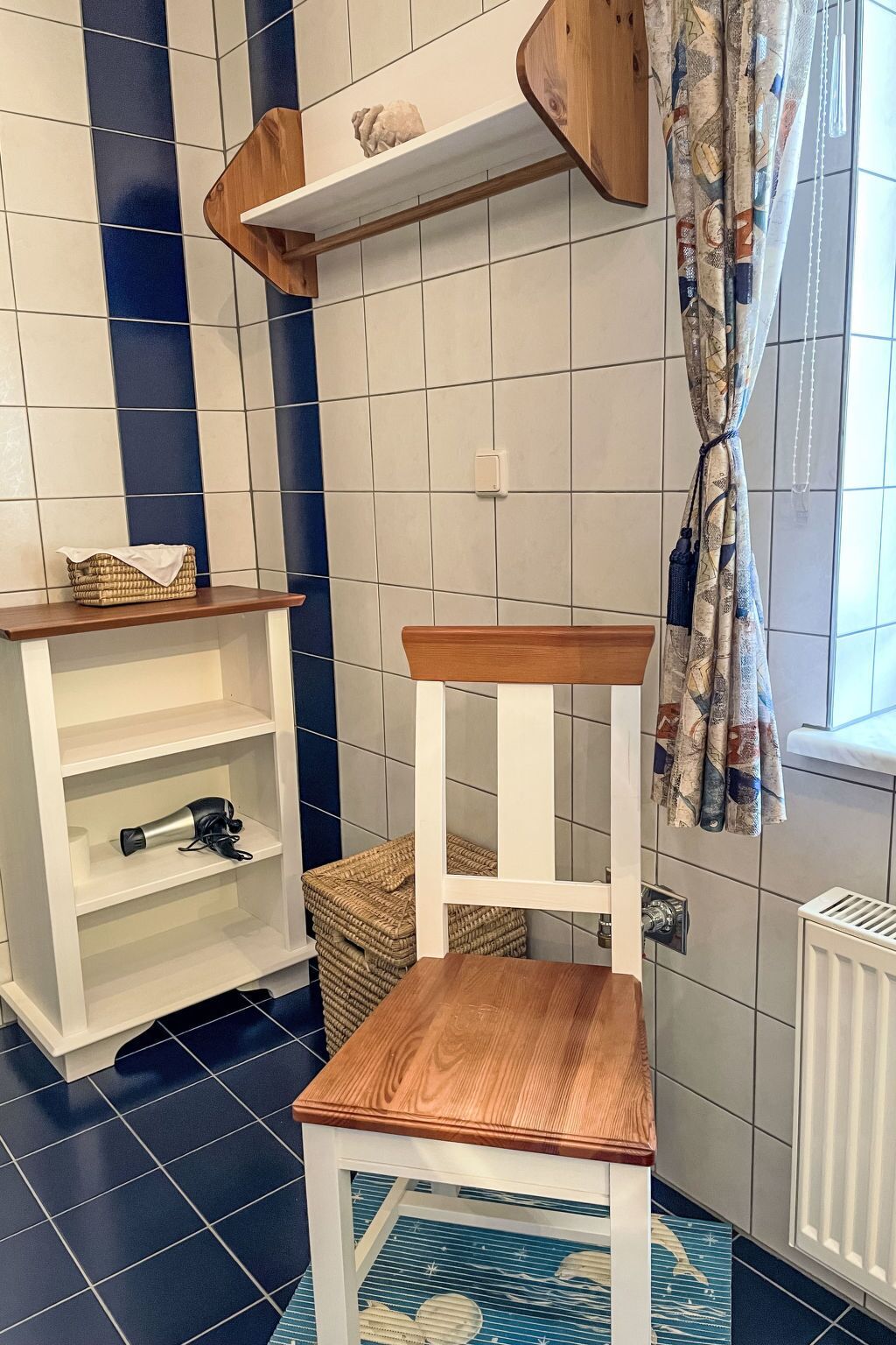 A bathroom with a white tiled wall, a wooden shelf with a decorative item, a white shelf with a hairdryer, a wooden chair, and a patterned rug.
