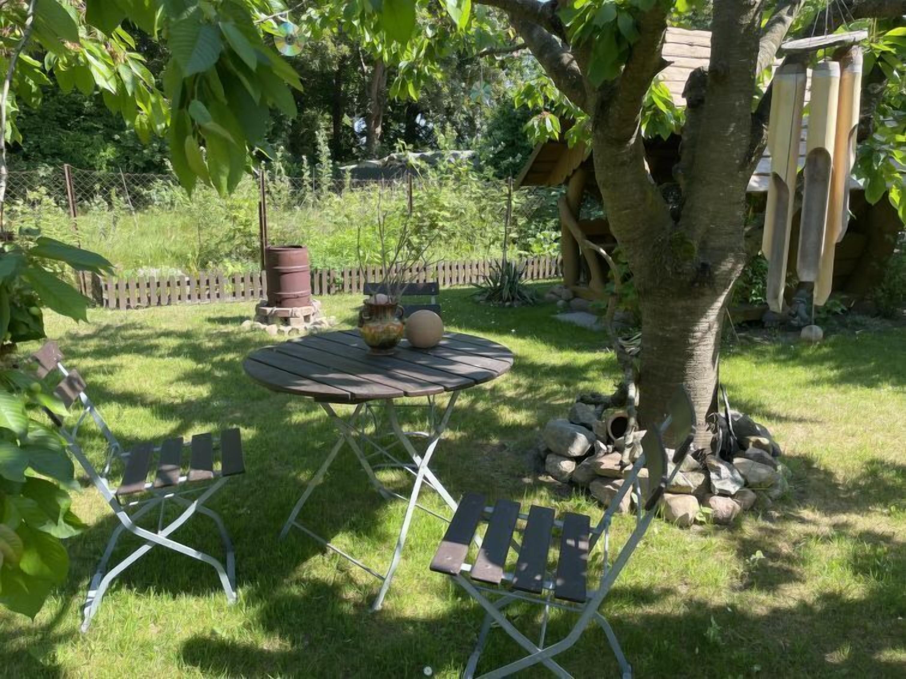 Una mesa redonda de madera con dos sillas de metal, una planta en maceta y un tronco de árbol en un espacio al aire libre con césped.