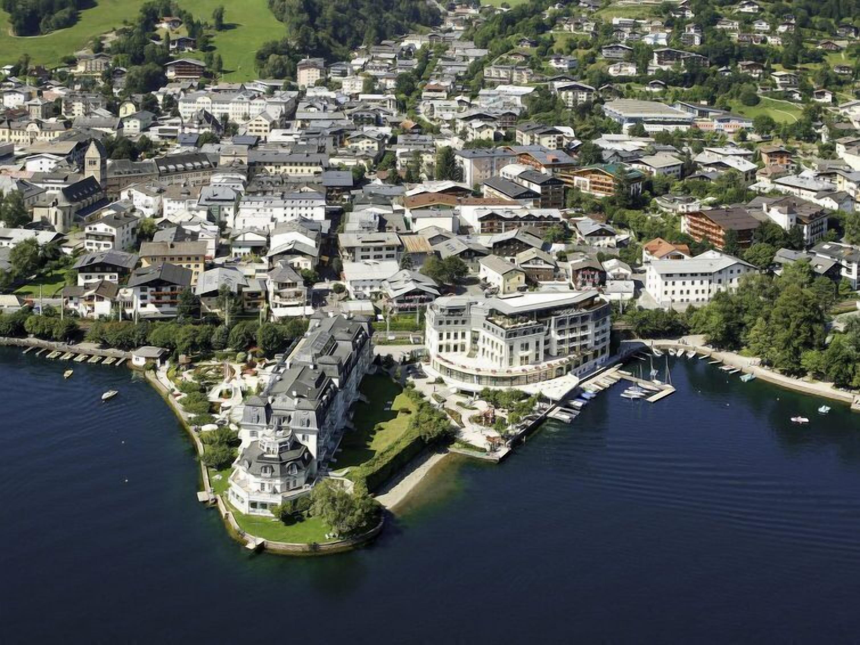 An aerial view of a coastal town with a prominent waterfront property featuring a large, multi-story building with a curved facade, a dock, and a marina with several boats.