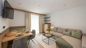 Una sala de estar moderna con un sofá, una mesa de comedor con sillas, un televisor montado en la pared y una cocina al fondo.