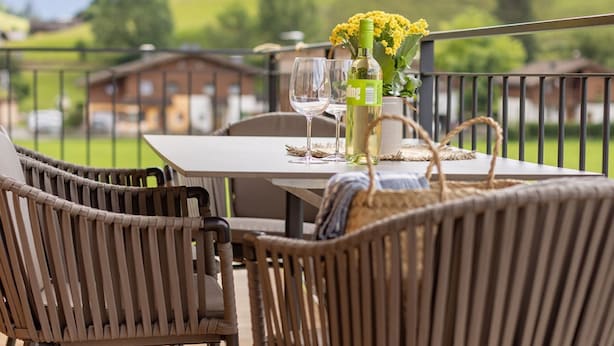 A dining table with chairs, a bottle of wine, and a vase of yellow flowers on a balcony with a mountain view.