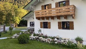 Un edificio de dos pisos con fachada blanca, balcón de madera y un muro de piedra al frente.