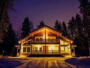 A two-story log cabin with a balcony, surrounded by snow and trees.
