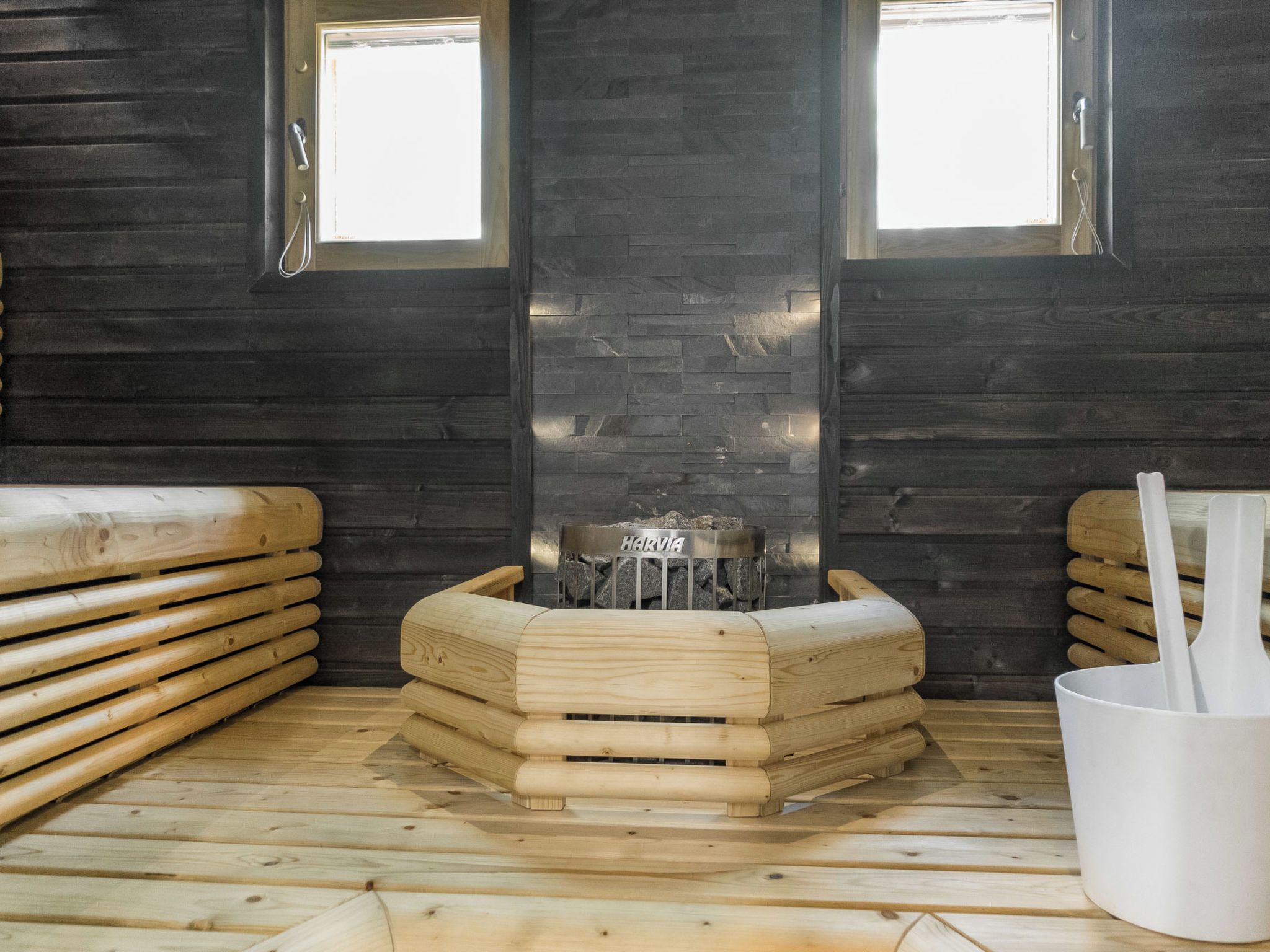 A sauna room with wooden benches, a central stone heater, and two windows.