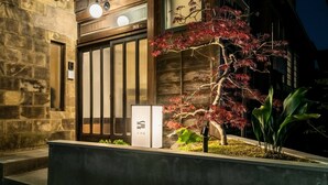 A modern building with a stone facade, a large wooden door, and a small garden with a red-leaved tree. - Sansuikyo Machiya House (Kanazawa)