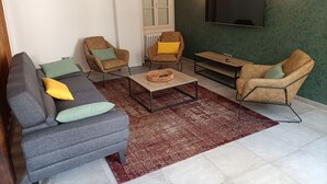 Ein Wohnzimmer mit grauem Sofa, zwei Sesseln, einem Couchtisch und einem Fernseher, der an einer grünen Wand befestigt ist.