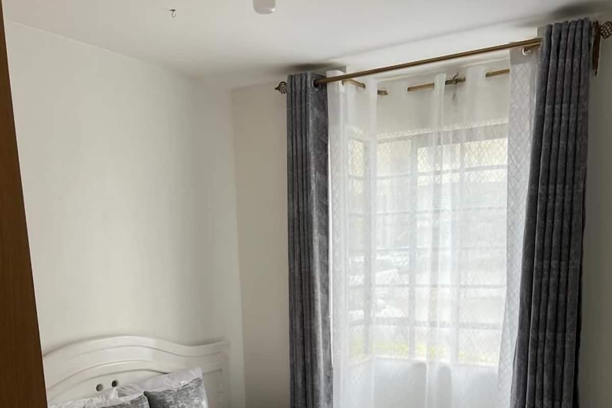 A neatly made bed with a white headboard, grey curtains, and a window with sheer white curtains.
