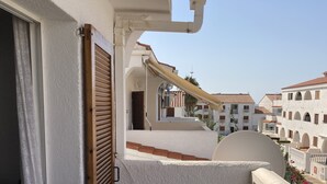 Un balcón con una pared blanca con textura, un persiano de madera y vistas a edificios vecinos con balcones y tejados de tejas.