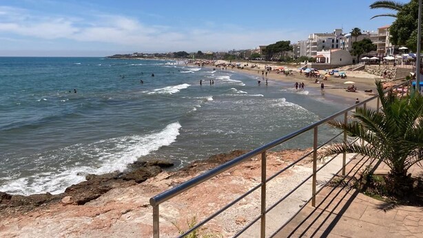 Escena costera de una playa con costa rocosa, un paseo marítimo y edificios al fondo.