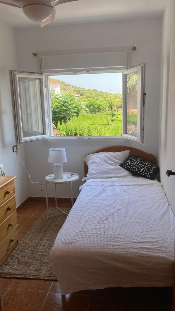 Een slaapkamer met een bed, een nachtkastje met lamp, een raam met uitzicht op groen en een houten dressoir.